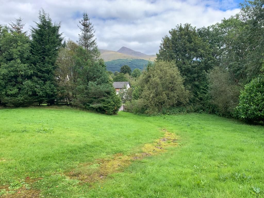 A grassy field with a path leading to a house surrounded by trees and hills in the background.
