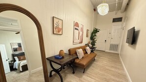 Un pasillo con piso de madera, un sofá marrón con cojines, una mesa de madera con un jarrón y un objeto azul, y un espejo grande.