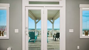 En vy genom en glasdörr ut mot en träveranda med stolar, med utsikt över en strand med strandhus.