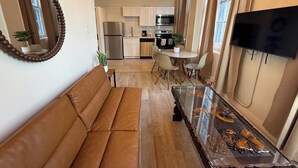 A modern living room with a brown leather sofa, a glass coffee table with decorative items, a dining area with a white table and chairs, a kitchen with white cabinets and stainless steel appliances, and a TV mounted on the wall.
