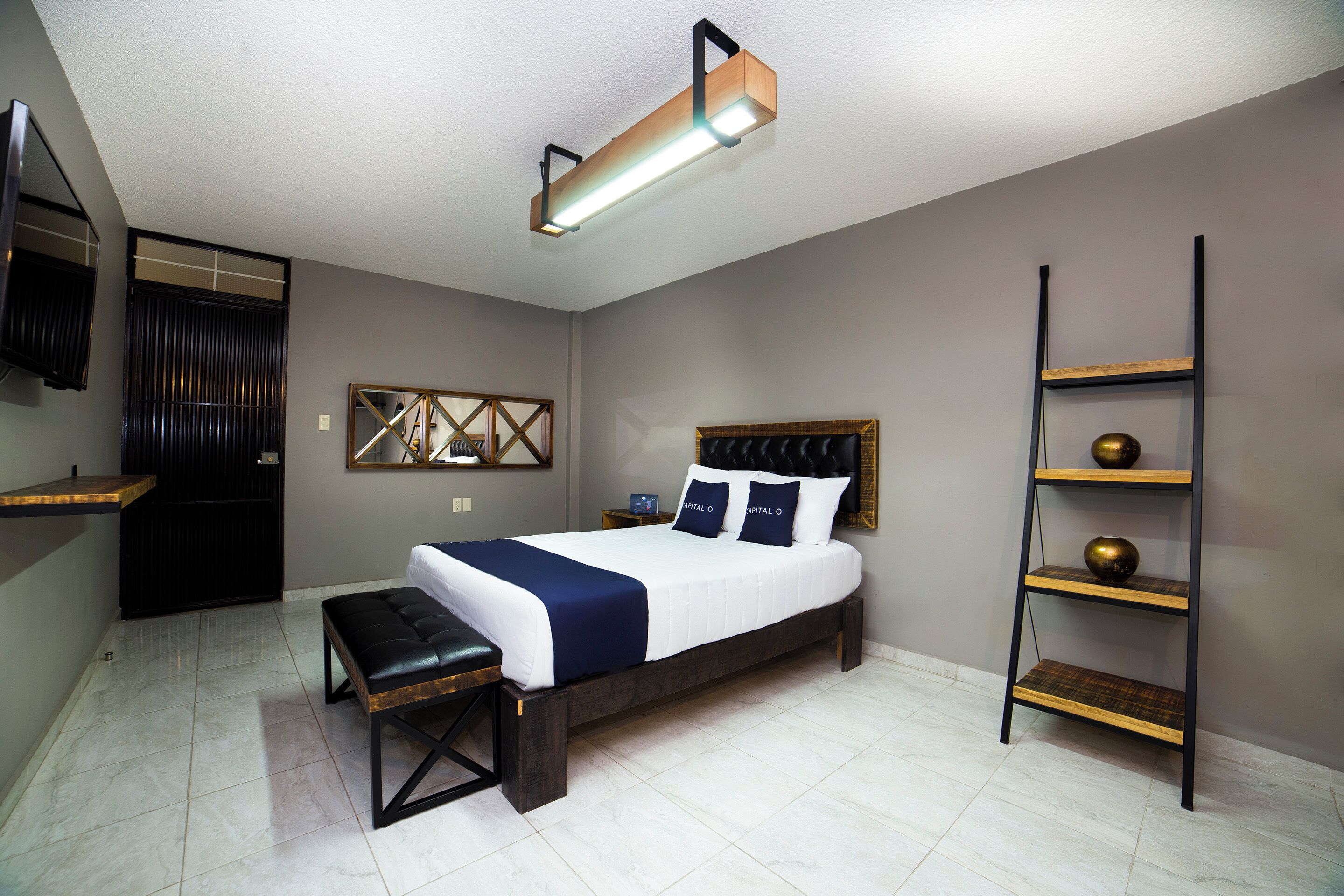 A bedroom with a bed, bench, bedside table, wall-mounted TV, and a shelf with decorative items.
