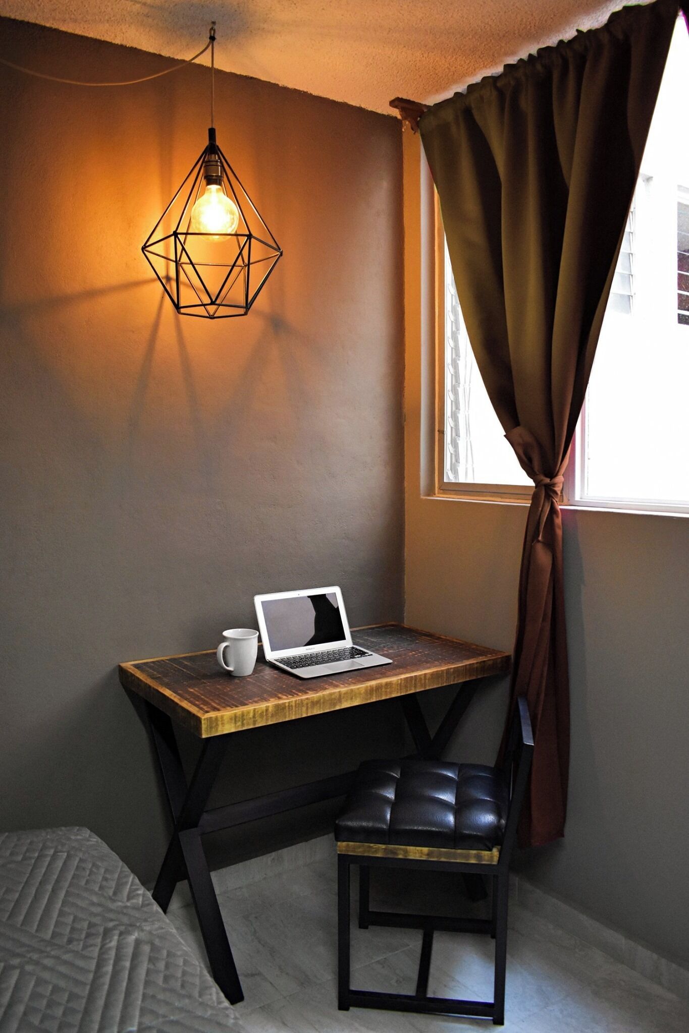 A corner room with a wooden desk, a laptop, a white mug, a black leather chair, a hanging light fixture, and a window with curtains.