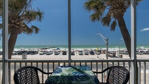 A view from a covered area with a table and chairs, overlooking a beach with parked cars and palm trees. - Beach View - Pelican Cove 3 by SeaBreeze Vacation (Bradenton Beach)