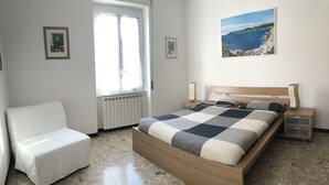 A bedroom with a bed, a chair, a nightstand, a lamp, and a painting of a coastal scene. - Sant'Antonino Apartments - Scirocco (levanto)