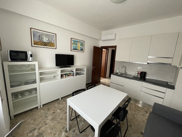 Cocina compacta con gabinetes blancos, microondas, televisor de pantalla plana y comedor con mesa y sillas.
