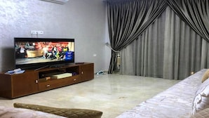 A living room with a flat-screen TV on a wooden stand, a bed with patterned bedding, and a large window with curtains. - Residence Hivernage Founty 4748 (Agadir)