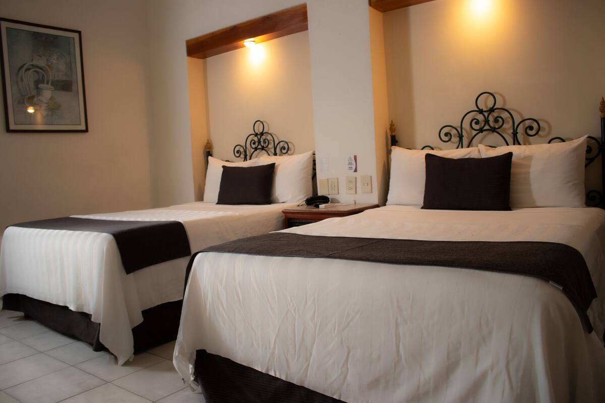 Two double beds with black and white bedding, a nightstand, a wall-mounted light, and a framed picture on the wall.