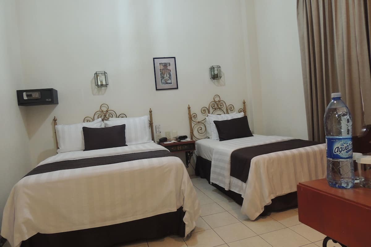 A hotel room with two beds, a bedside table, a nightstand, a wall-mounted lamp, and a framed picture.