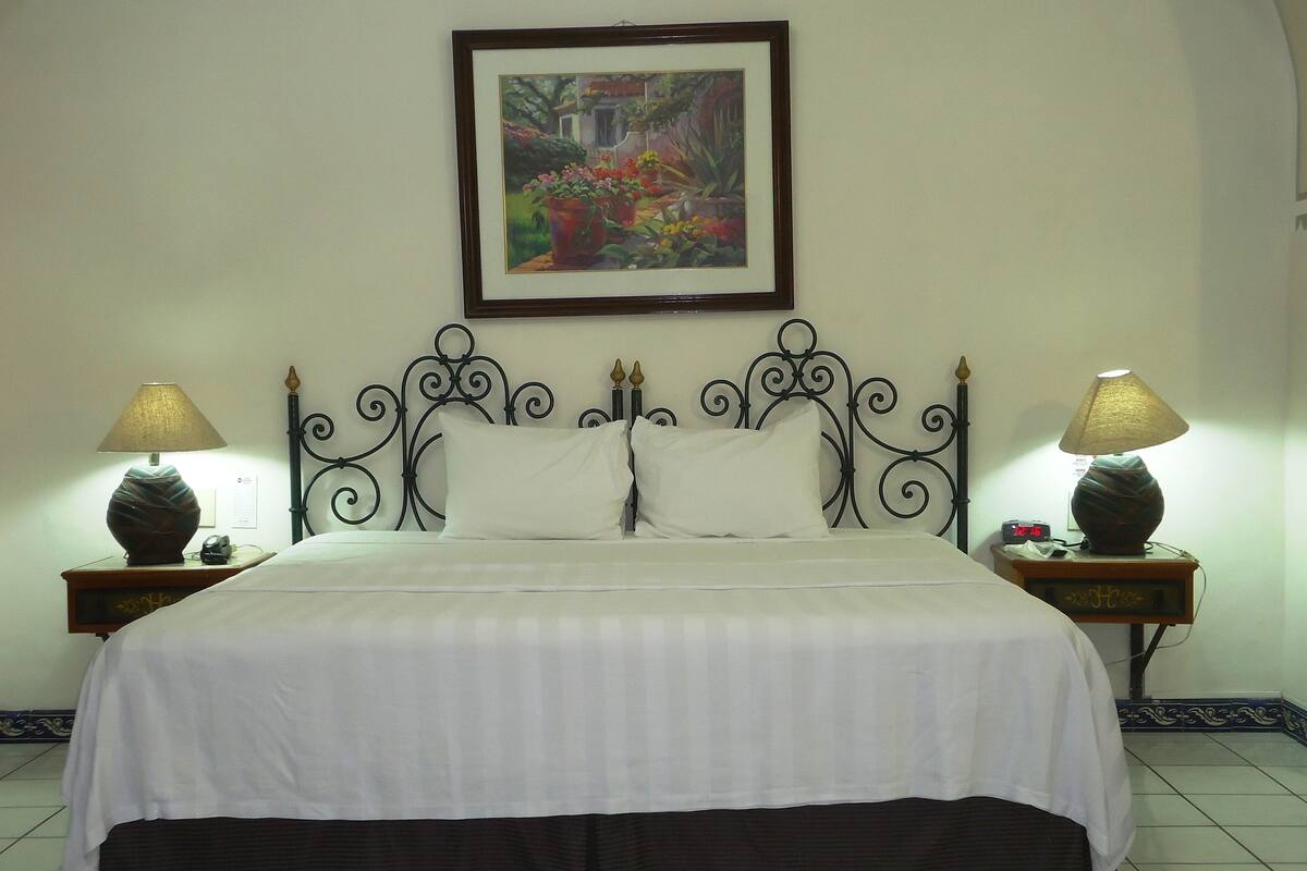 A neatly made bed with white linens, two bedside lamps, a framed painting above the bed, and a small clock on the nightstand.