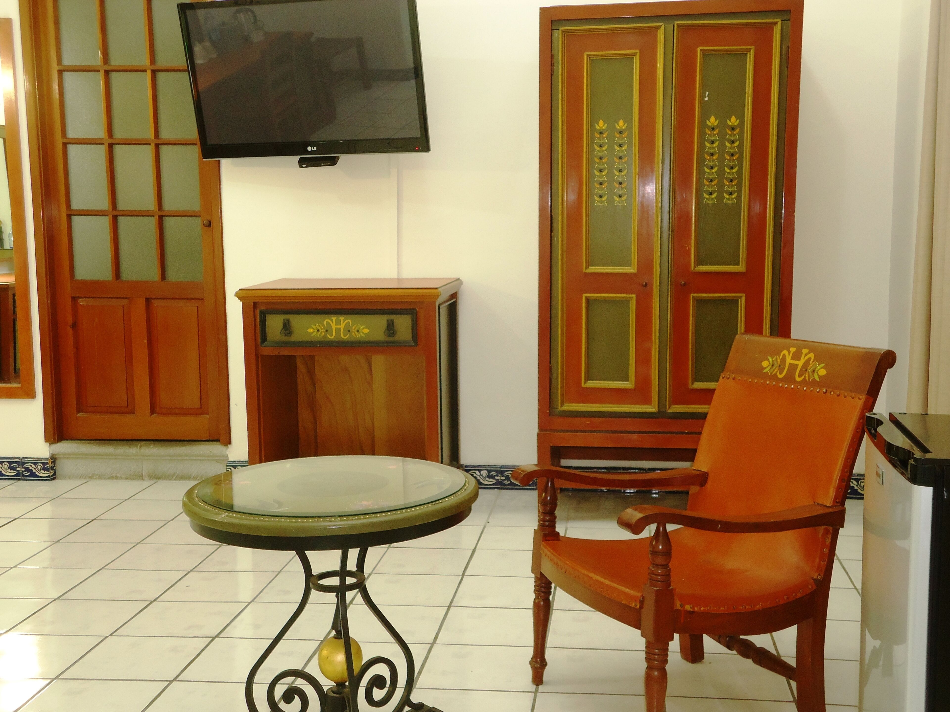 Quarto com televisão na parede, armário de madeira com portas de vidro, mesa redonda com tampo de vidro e cadeira laranja com encosto decorativo.