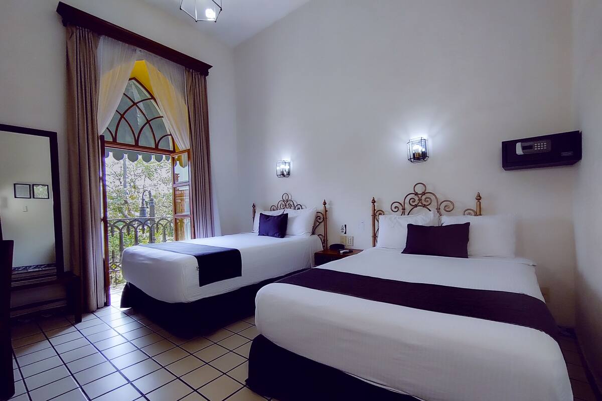 A hotel room with two beds, a balcony with curtains, and a view of greenery.
