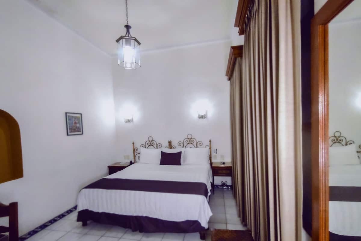 A hotel room with a bed, bedside tables, a hanging light fixture, a mirror, and a door with curtains.