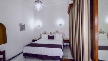 A hotel room with a bed, bedside tables, a hanging light fixture, a mirror, and a door with curtains.