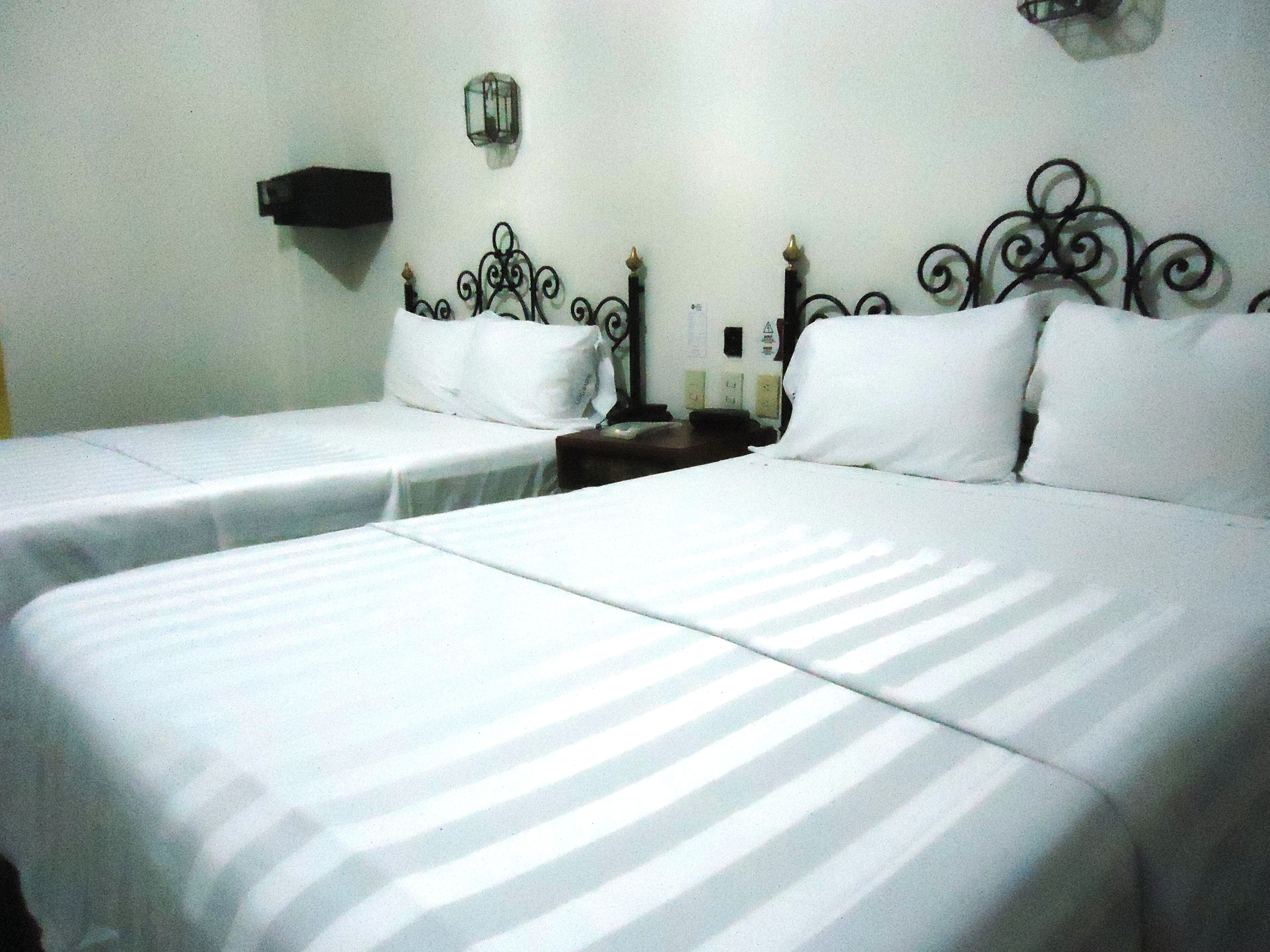 A hotel room with two beds, each with white bedding and a metal headboard. There is a nightstand with a lamp and a small table between the beds.