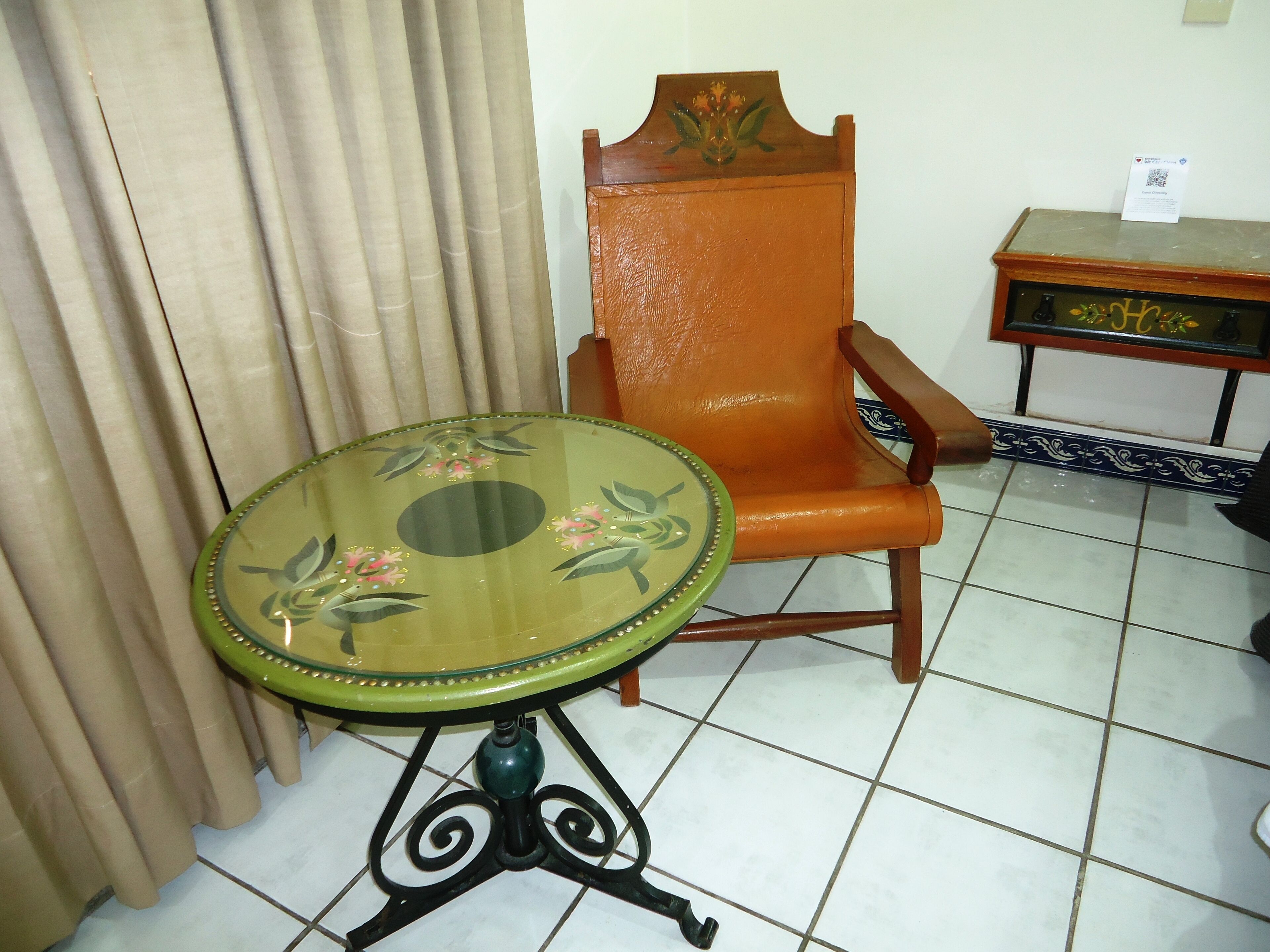 Una habitación con una mesa verde con motivos florales, una silla naranja y un pequeño armario de madera con un adorno en la parte superior.