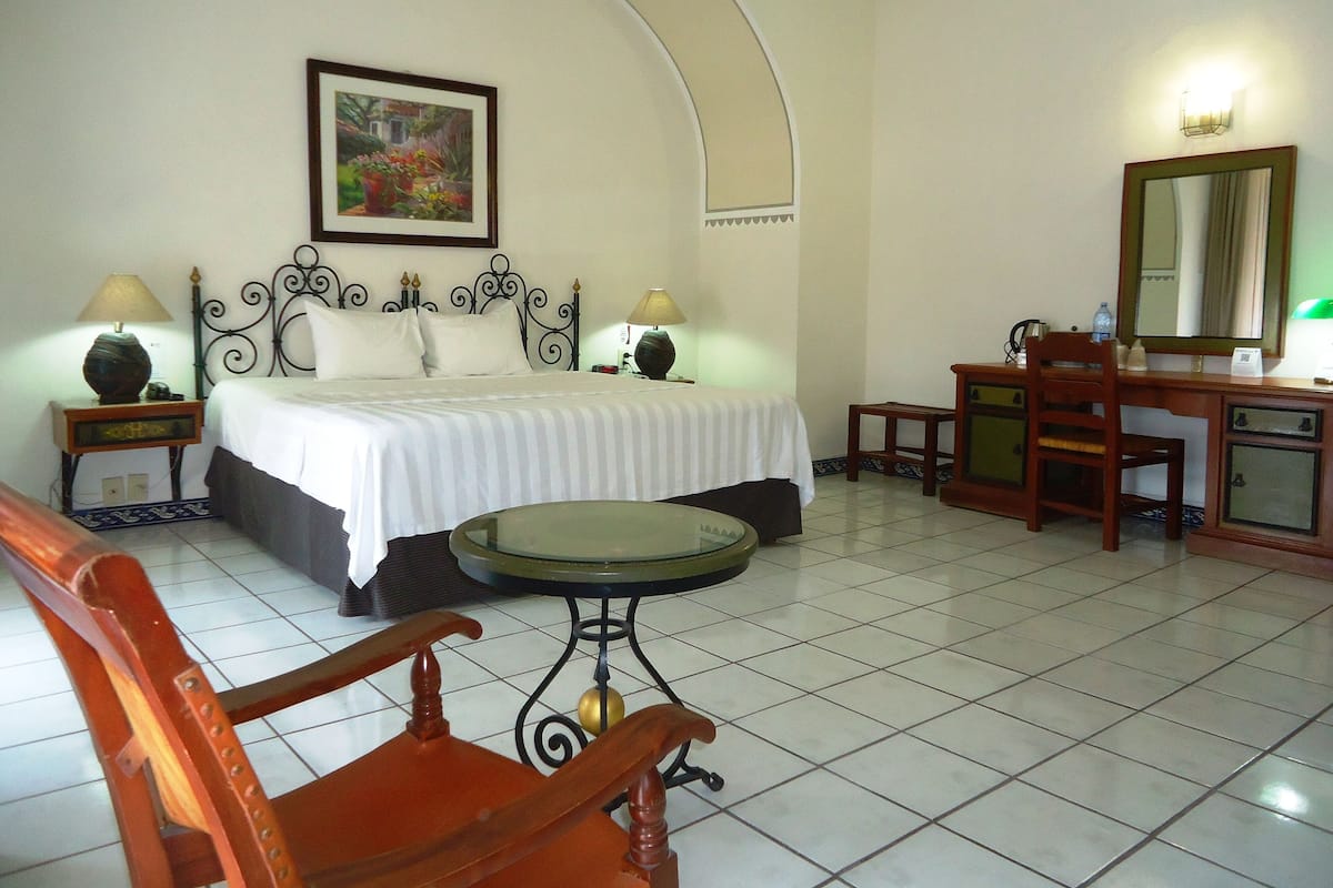 A hotel room with a bed, a desk, a chair, a table, and a painting on the wall.
