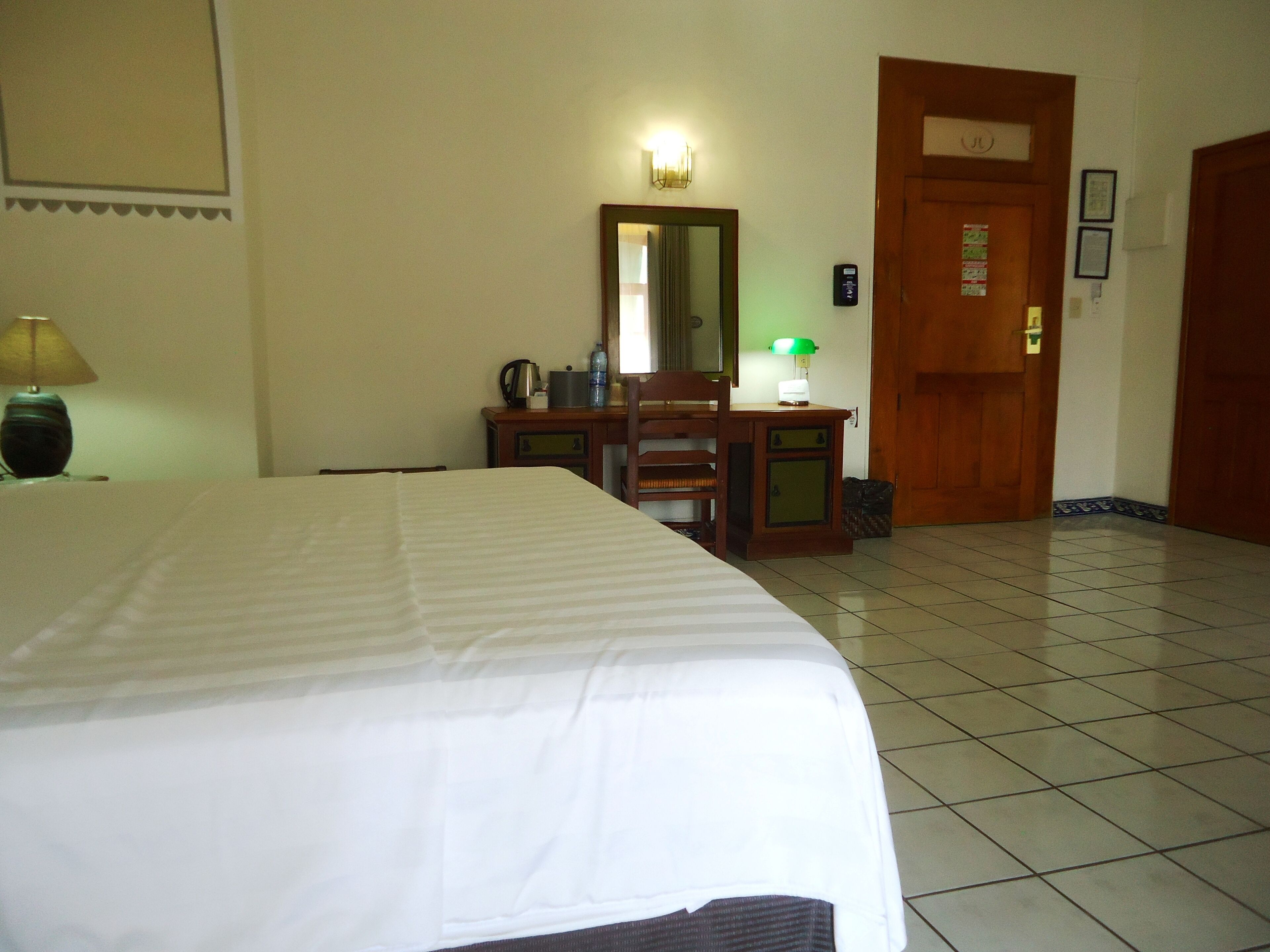 A hotel room with a bed, a desk with a chair, a mirror, a lamp, and a door.