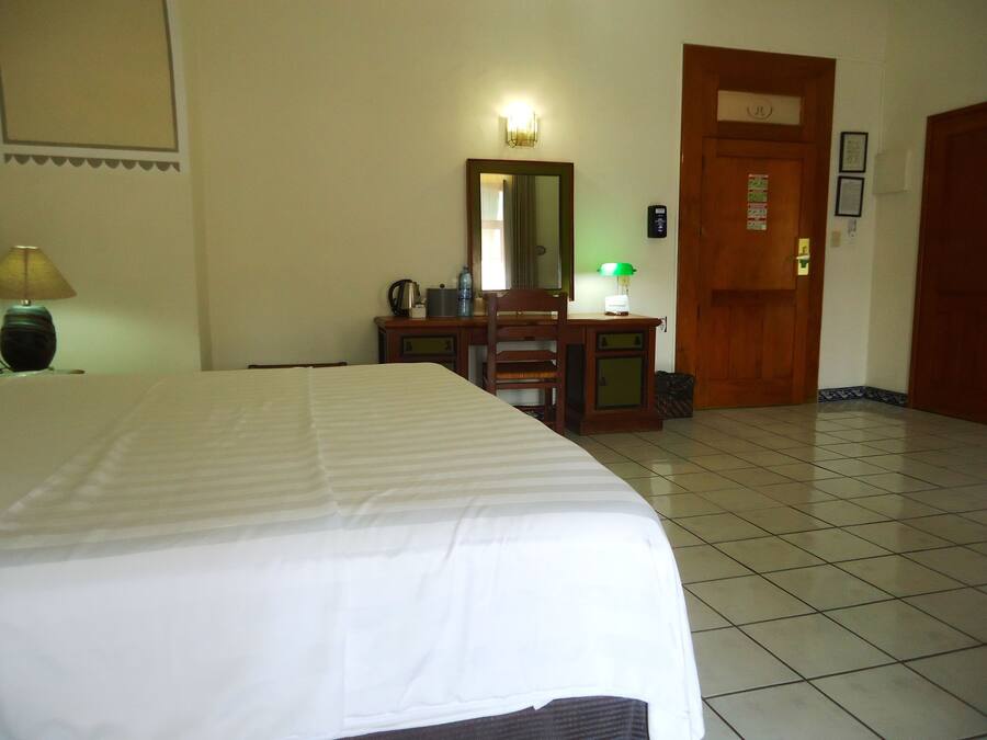 A hotel room with a bed, a desk with a chair, a mirror, a lamp, and a door.