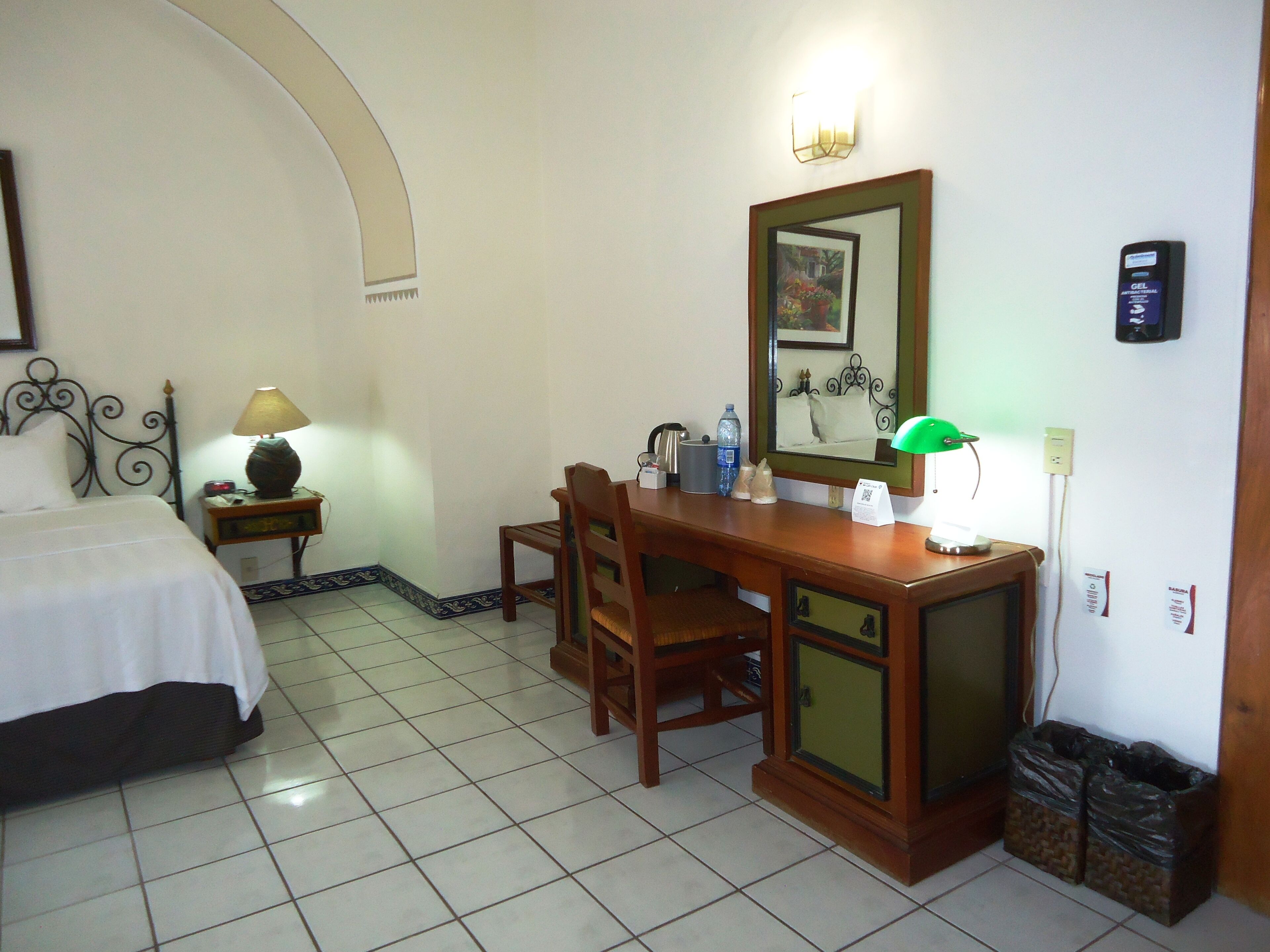 A hotel room with a bed, a desk, a chair, a mirror, a lamp, a trash bin, and a wall-mounted power outlet.