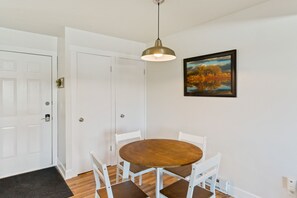 A dining area with a round wooden table and white chairs, a pendant light, and a framed painting on the wall. - Mountainside Condo: King Bed/Hot Tub/On Free Bus Route/Fireplace (Steamboat Springs)