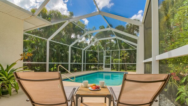 A covered outdoor area with a pool, two chairs, a table with fruit and cups, and lush greenery in the background. - Private Heated Pool & Beach House: 3 King Beds , TVs throughout, 1000mbps WiFi (Vero Beach)