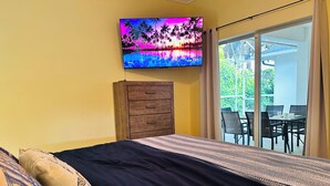 A bedroom with a bed, a wooden dresser, a TV on the wall, and a sliding glass door leading to an outdoor area with a table and chairs. - Private Heated Pool & Beach House: 3 King Beds , TVs throughout, 1000mbps WiFi (Vero Beach)