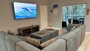 A living room with a grey sectional sofa, a flat-screen TV displaying dolphins, a wooden coffee table, and a sliding glass door leading to a patio. - Private Heated Pool & Beach House: 3 King Beds , TVs throughout, 1000mbps WiFi (Vero Beach)