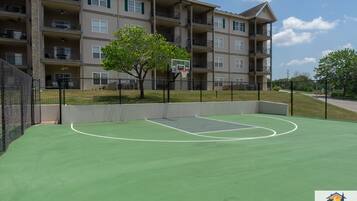 Un terrain de basket avec un panier, entouré d’une clôture, et un immeuble à plusieurs étages en arrière-plan.