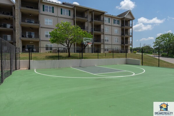 A basketball court with a net, surrounded by a fence, and a multi-story building in the background.