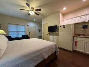 A hotel room with a bed, a ceiling fan, a television, a refrigerator, and a small kitchen area.