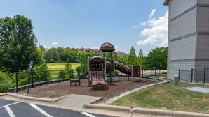 A playground with a slide and climbing structures, surrounded by a fence and a building in the background.