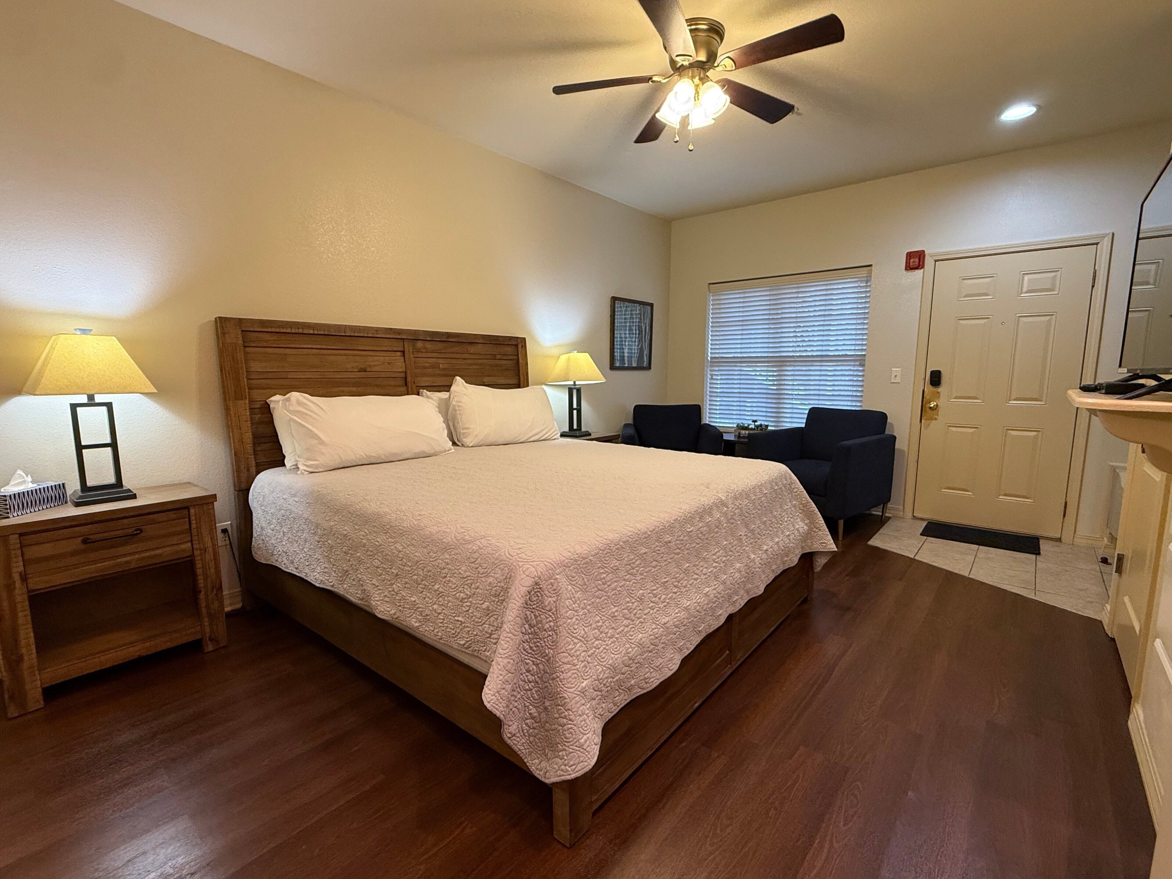 A hotel room with a bed, two bedside lamps, a ceiling fan, a door, and a window with blinds.