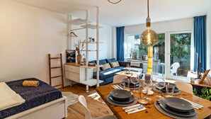 A modern living room with a sofa, dining table set for four, and a staircase with a white railing. - Cosy Studio Apartment in Quiet Area with Wi-Fi, Terrace & Garden (Pforzheim)