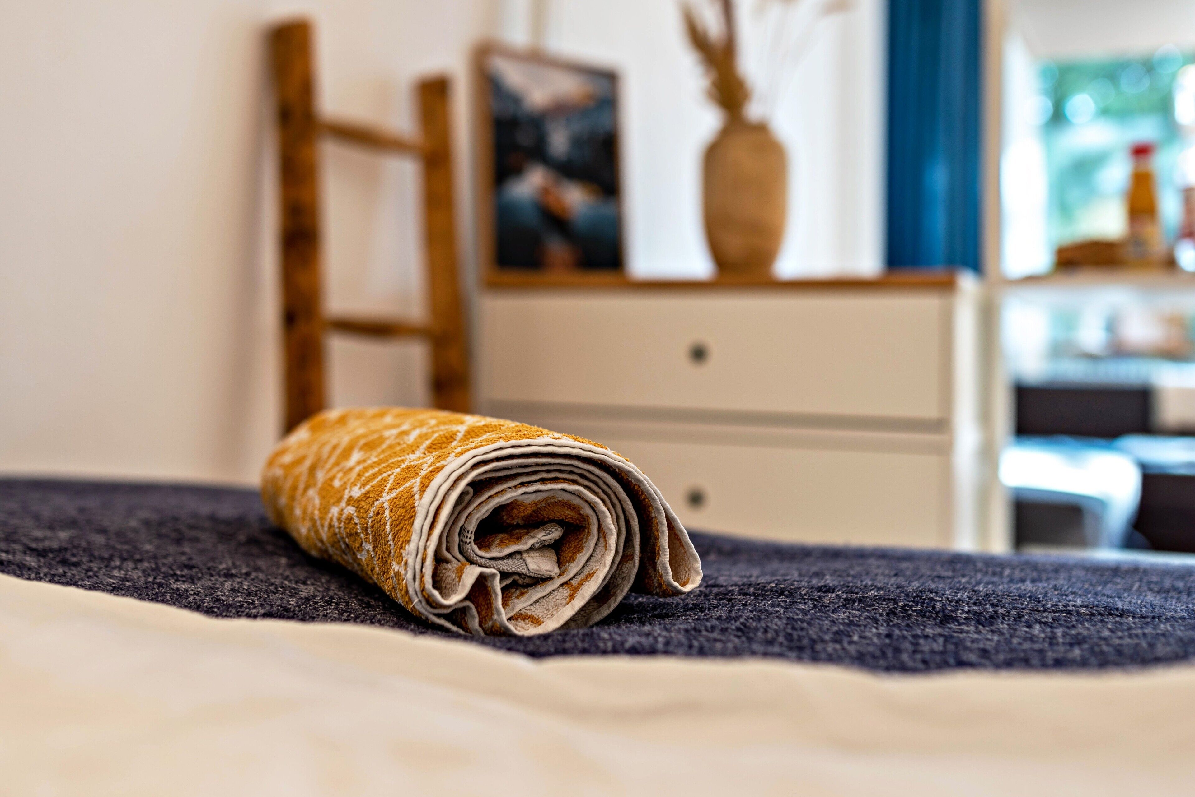 Een netjes opgemaakt bed met een opgerolde handdoek, een houten stoel, een dressoir en een ingelijst beeld aan de muur.