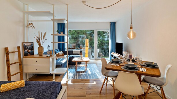 A modern, well-lit room with a dining area, a bed with a blue headboard, and a view through large windows. - Cosy Studio Apartment in Quiet Area with Wi-Fi, Terrace & Garden (Pforzheim)
