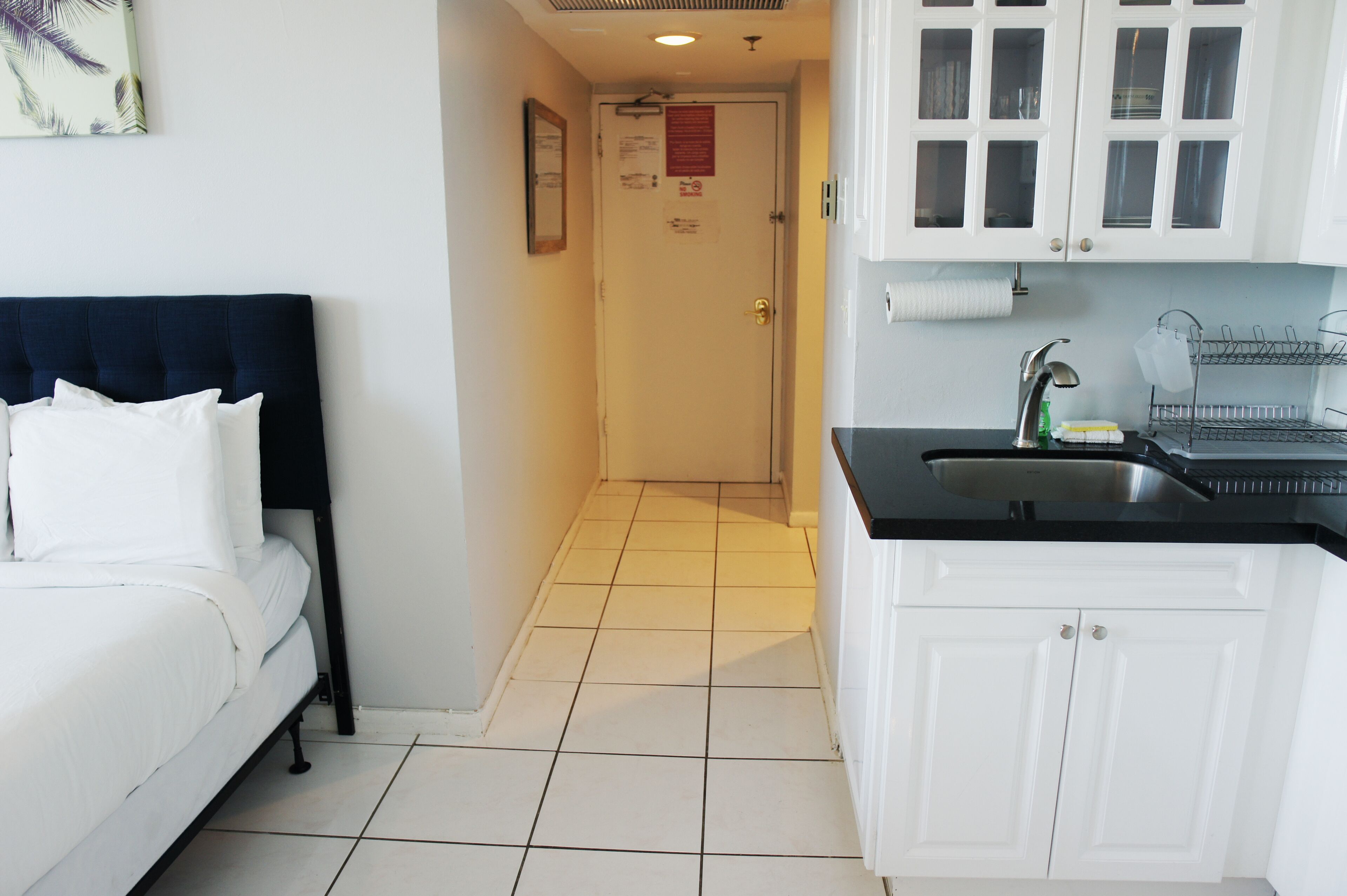 Une chambre d’hôtel avec un lit, un coin cuisine doté de meubles blancs, un plan de travail noir et un sol carrelé.