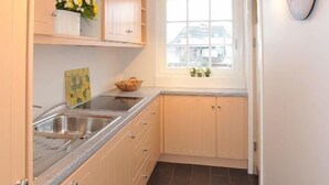 A narrow kitchen with light wooden cabinets, a window with a view, and a clock on the wall. - Biberhüs OG (Sylt)