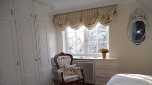 A bedroom with a large window, a radiator, a chair with a bird pattern, and a bed with a geometric quilt. - Biberhüs OG (Sylt)