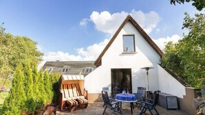 Une maison blanche avec un toit en pente, une terrasse avec des meubles d’extérieur et un petit jardin.