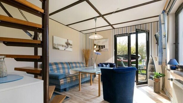 Un salon moderne avec un canapé, une table basse et un fauteuil bleu. On y trouve un escalier avec des rampes en bois et une vue sur l’extérieur grâce à de grandes fenêtres.