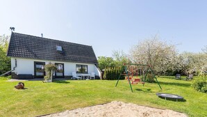 Une maison avec un toit en tuiles, une terrasse avec des chaises, une balançoire et un trampoline dans le jardin.
