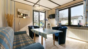 Un salon confortable avec un canapé, une table et des chaises. Un téléviseur est fixé au mur et une fenêtre offre une vue sur l’extérieur.