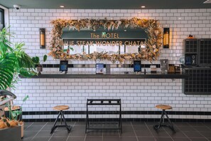Un espace dâaccueil dâhĂŽtel avec un mur recouvert de carreaux blancs, une guirlande de fleurs au-dessus du comptoir dâaccueil, une horloge numĂ©rique et deux tabourets.