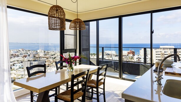 Une cuisine moderne dotée d’une grande fenêtre, un coin repas avec une table et des chaises, et une vue sur la ville et la mer.