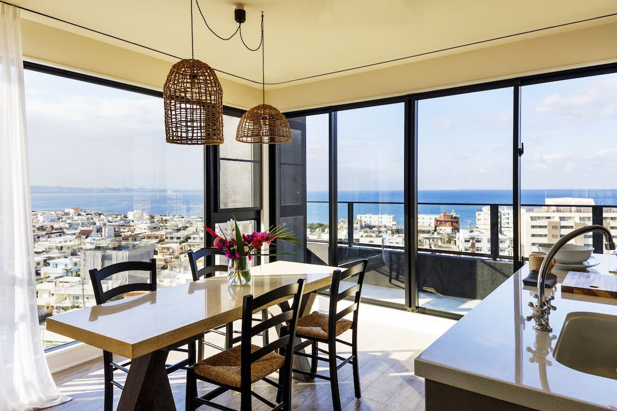 Une cuisine moderne dotée d’une grande fenêtre, un coin repas avec une table et des chaises, et une vue sur la ville et la mer.