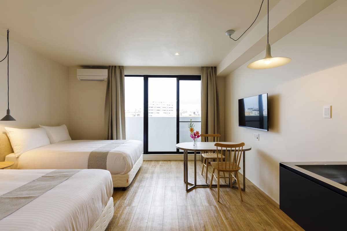 Une chambre d’hôtel avec deux lits, une table à manger avec des chaises, une télévision et une grande fenêtre avec des rideaux.