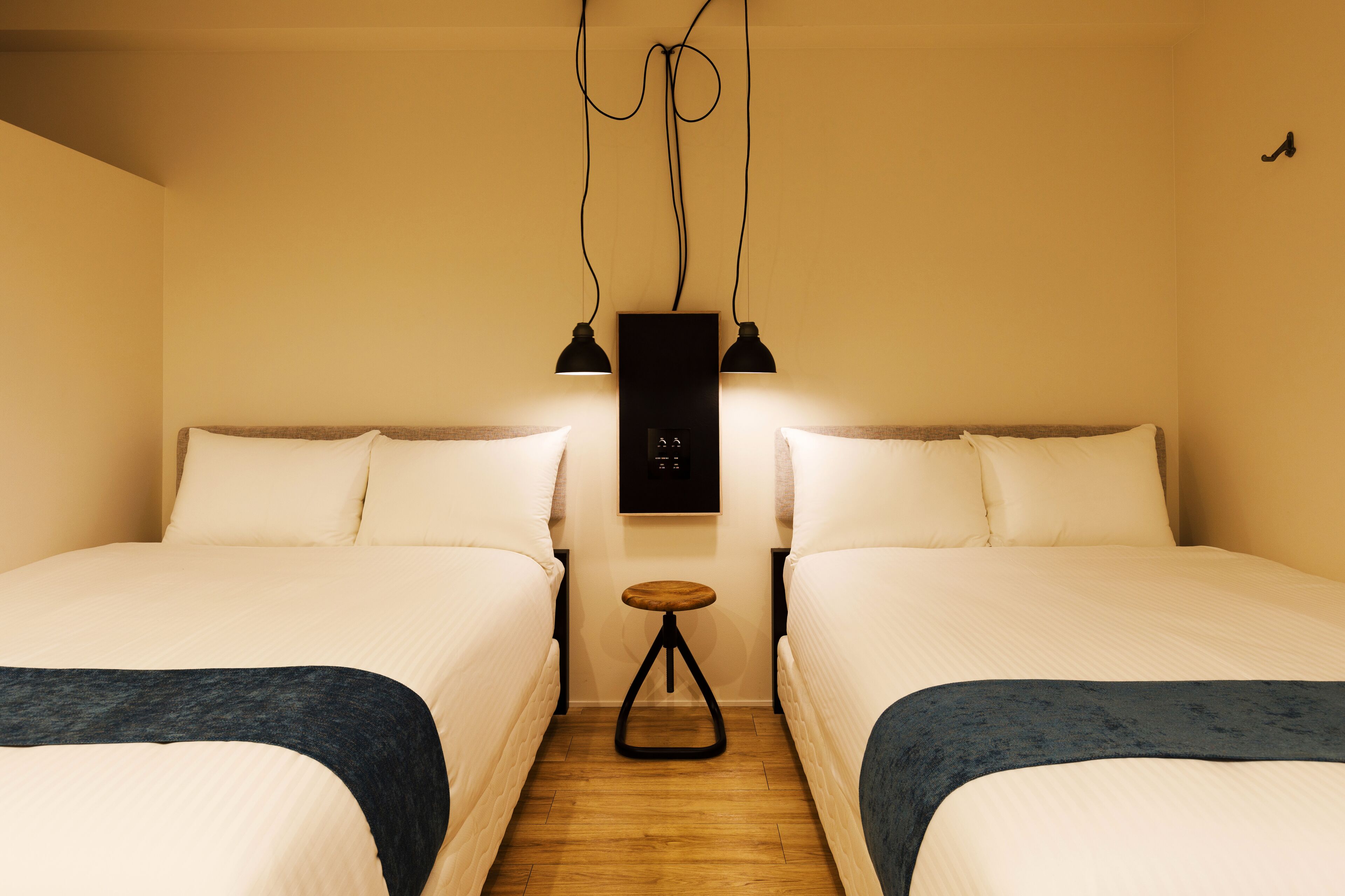 A hotel room with two beds, a small wooden stool, and a wall-mounted control panel.