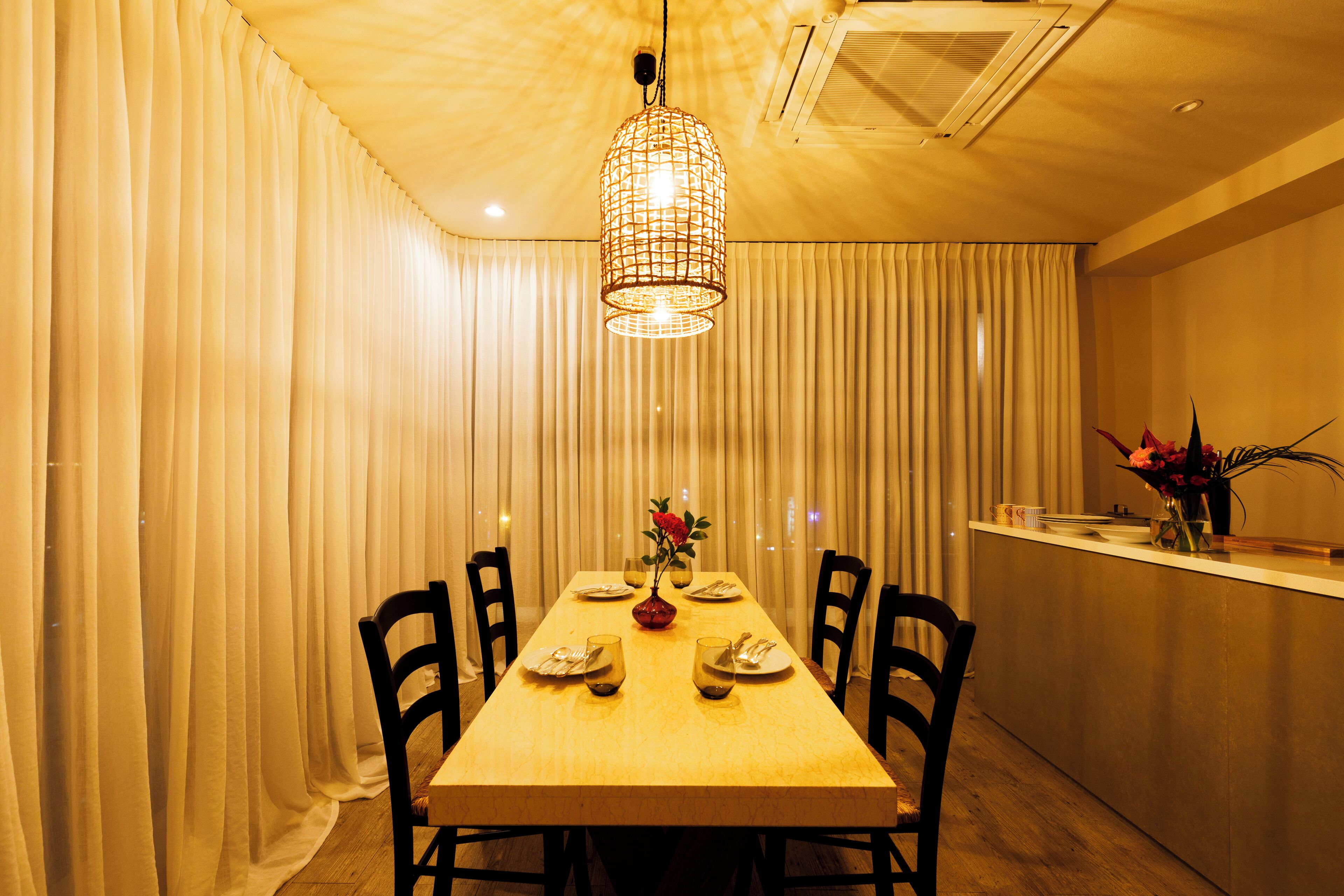 A dining area with a wooden table set for four, surrounded by chairs, a hanging light fixture, and a counter with a vase of flowers.