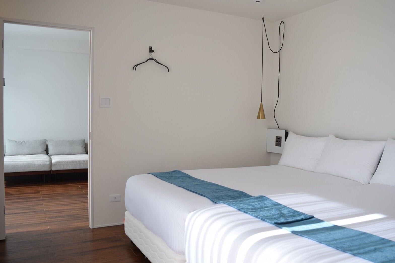 A bedroom with a bed, a hanging light, a door, and a sofa in the adjacent room.