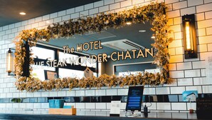 Un espace d’accueil agrémenté d’un miroir encadré de fleurs séchées, d’une enseigne indiquant « THE HOTEL BLUE STEAK WONDER CHATAN » et d’un écran numérique affichant un message de bienvenue.
